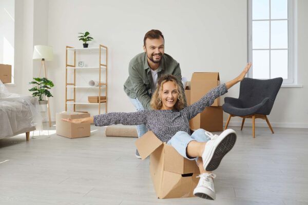 Two young people buy house and make dream come true. Happy married couple having fun in new home on moving day. Joyful first time buyers playing with boxes in modern living room. Real estate concept