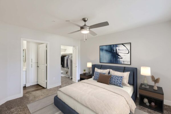 Pavilions at Arrowhead apartment bedroom with carpet, plush bed with headboard, bedside tables with lamps, and ceiling fan.