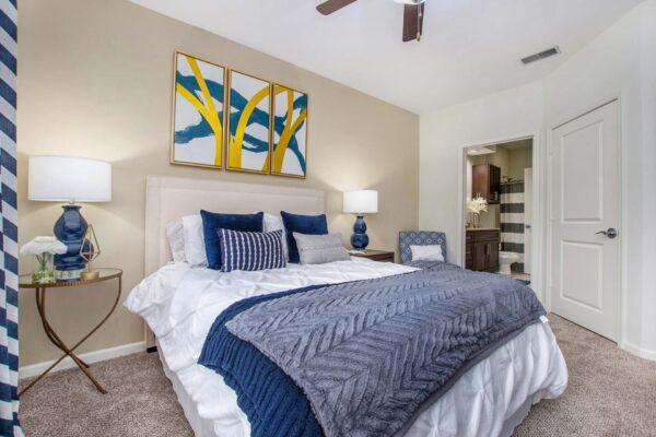 Village at Mission Farms apartment bedroom with carpet, plush bed with headboard, bedside tables with lamps, and framed artwork.