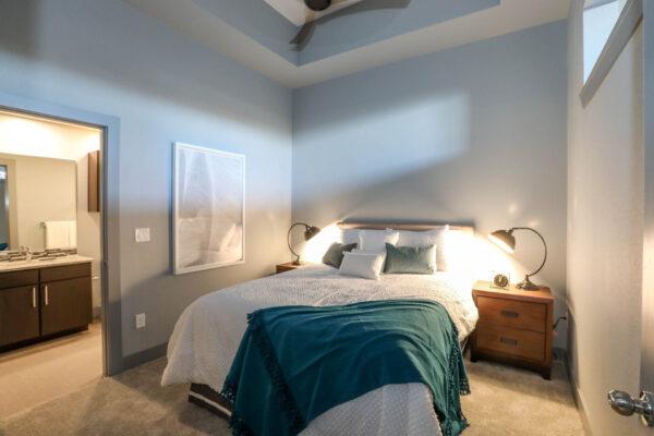 Forum Fitzsimons apartment bedroom with carpet, plush bed with headboard, and bedside tables with lamps.