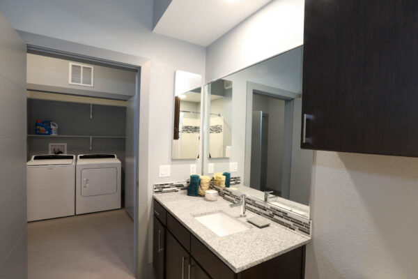 Forum Fitzsimons apartment bathroom with dark cabinets, light counters, large mirror, and door to laundry machines.