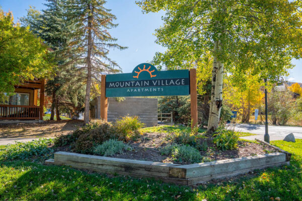 Mountain Village apartments building sign in grassy area with big trees.