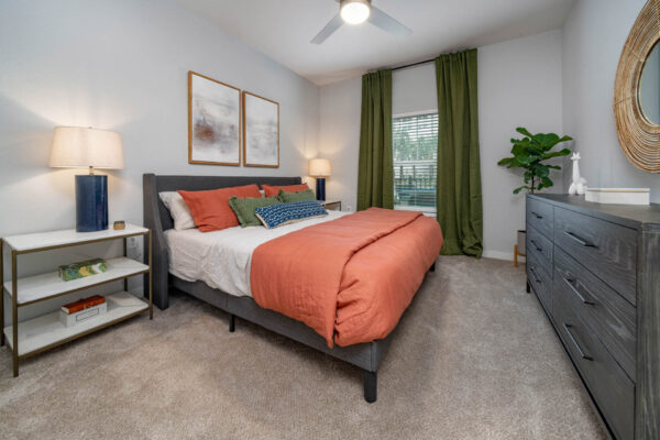 The Avery apartment bedroom with carpet, plush bed with headboard, bedside tables with lamps, and large window.