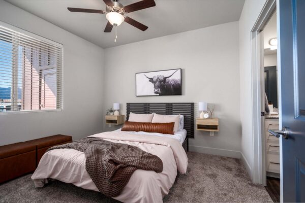 The Flats at Aero Park apartment bedroom with carpet, plush bed with headboard, ceiling fan, and large window.